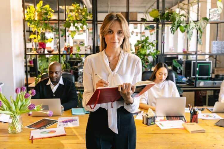 Photo by ANTONI SHKRABA from Pexels: https://www.pexels.com/photo/woman-standing-with-a-notebook-and-people-working-on-laptops-behind-her-in-an-office-7163456/